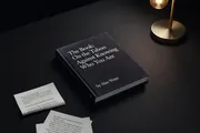 A black hardcover titled The Book: On the Taboo Against Knowing Who You Are, by Alan Watts, on a dark desk under a brass lamp, loose printed pages beside it