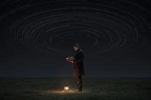 A person at the edge of a dark field at night holds a notebook under a long arc of turning stars, a warm lantern at their feet