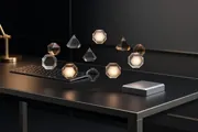 Agent Mode Changes the Shape of Thought blog hero. Translucent glass polyhedra glowing softly, floating in a row above a keyboard on a dark desk, the unit of thought lifted off the keys.