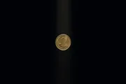 A single worn brass coin suspended in a shaft of warm light against deep black, its two faces caught between turns.