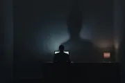 A lone figure at a desk in cold screen light, their shadow on the wall thinning and dissolving into darkness, a single warm lamp holding back the corner.