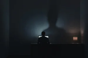 A lone figure at a desk in cold screen light, their shadow on the wall thinning and dissolving into darkness, a single warm lamp holding back the corner.