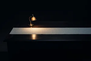 A long sheet of blank ruled ledger paper under a small warm desk lamp, most of its length fading into deep shadow.