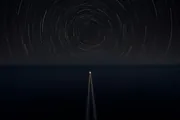 Star trails turning in closed circles over a dark sea, and below them the single straight wake of a small boat heading for the horizon