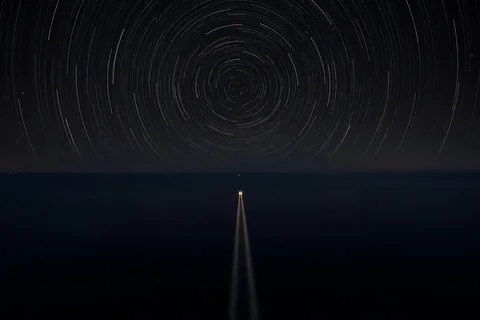 Star trails turning in closed circles over a dark sea, and below them the single straight wake of a small boat heading for the horizon
