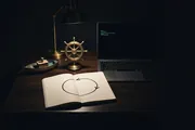 A small brass ship's wheel and a tin toy boat on a dark desk beside a laptop, a notebook open to one hand-drawn arrow that loops back on itself, lit by one warm lamp